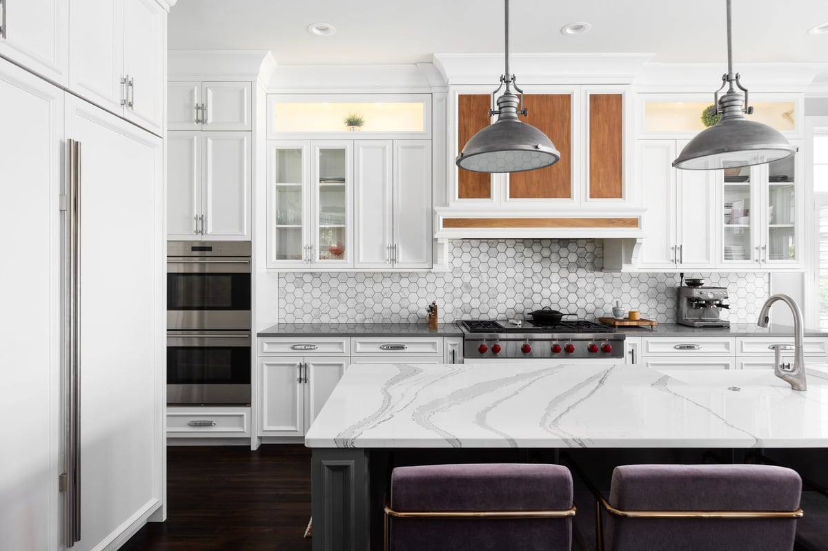 A kitchen detail with metal lights hanging above a large marble island, stainless faucet on a white farmhouse sink, and a marble hexagon tiled backsplash with white cabinets