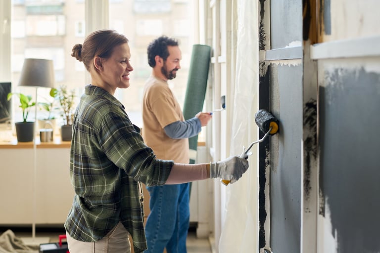 Side view of young happy woman with paintroller applying grey paint on wall of living room against her husband
