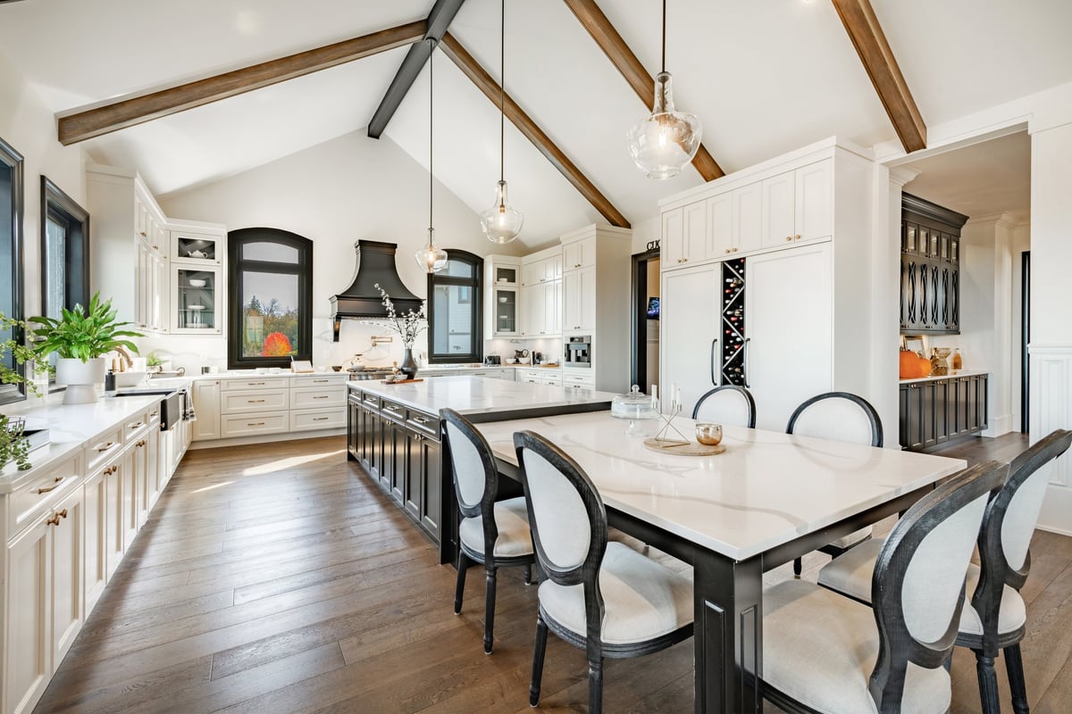 This is a huge kitchen with a modern farmhouse design style the countertops are white granite or marble and the kitchen island is very long and expansive with dining chairs there are houseplants cookbooks small appliances and staging decor on the counters. There is a large wine rack and stainless steel appliances. Hardwood floors wooden beams on the vaulted ceiling, the cabinets are a very dark wood and the windows are trimmed in black with a view to a bright blue sky autumn day showcasing a very colourful scene.